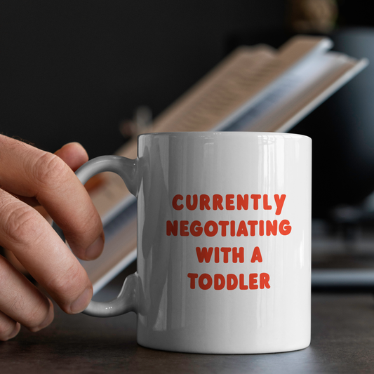 White mug with red text 'Currently Negotiating with a Toddler' held by a hand on a table.