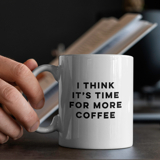 White mug with 'I think it's time for more coffee' text held by a hand on a blurred background