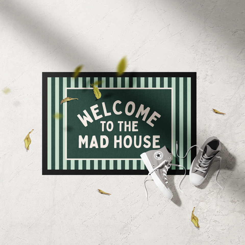 Doormat with 'Welcome to the Mad House' text on a white floor with shoes and leaves.