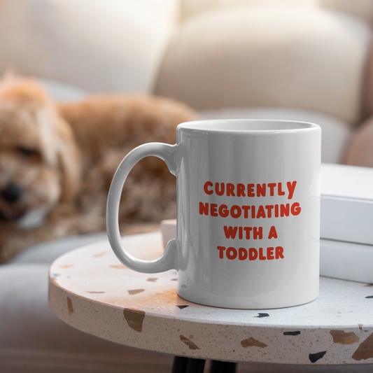 White mug with red text 'Currently negotiating with a toddler' on a small table with a dog in the background.