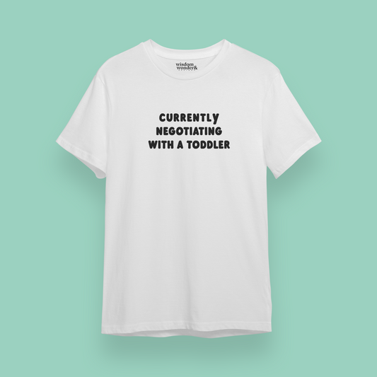 White t-shirt with text 'Currently Negotiating with a Toddler' on a light green background