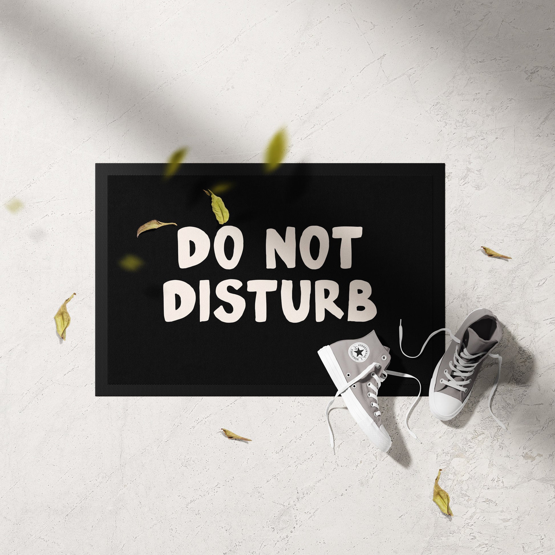 Doormat with 'DO NOT DISTURB' text on a light gray floor with white sneakers and yellow leaves.