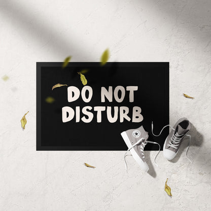 Doormat with 'DO NOT DISTURB' text on a light gray floor with white sneakers and yellow leaves.