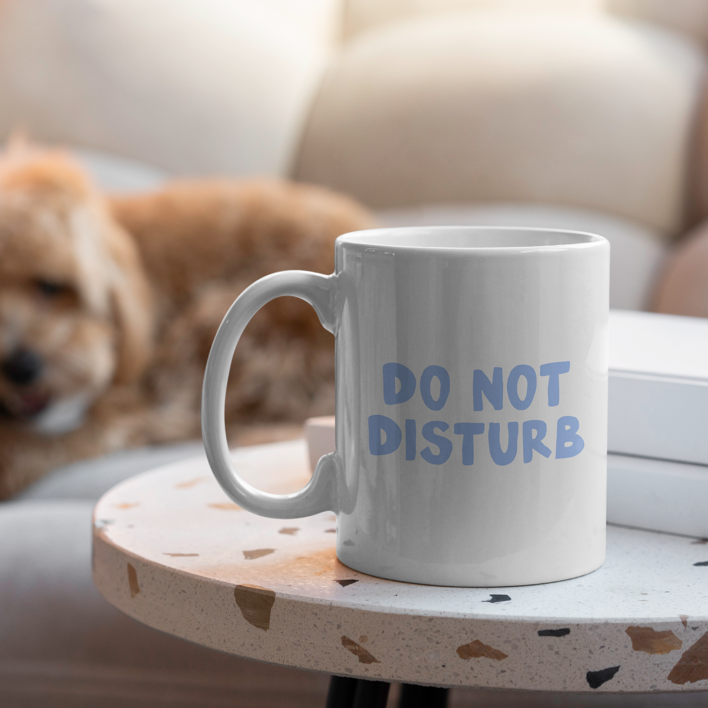 White mug with 'DO NOT DISTURB' text on a small table with a dog in the background.