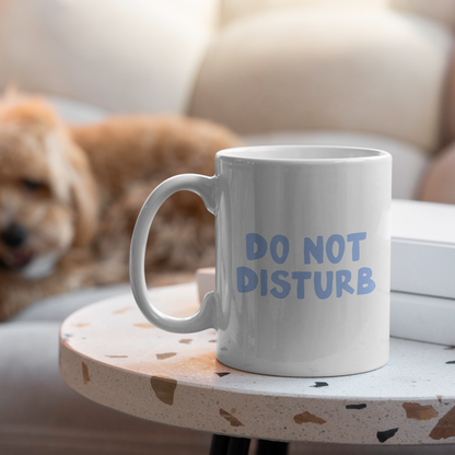 White mug with 'DO NOT DISTURB' text on a small table with a dog in the background.