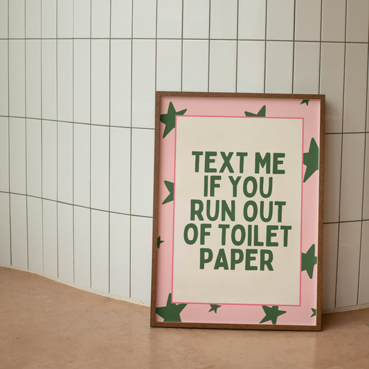 Framed poster with text 'Text me if you run out of toilet paper' on a tiled wall.