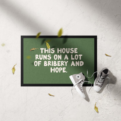 Doormat with text 'This house runs on a lot of bribery and hope' on a light gray floor.