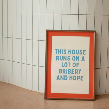 Framed poster with text 'This house runs on a lot of bribery and hope' against a tiled wall.