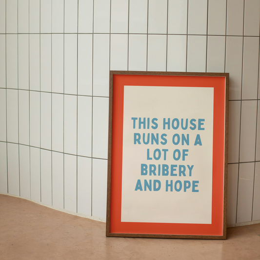 Framed poster with text 'This house runs on a lot of bribery and hope' against a tiled wall.