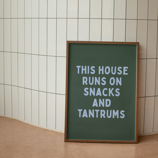 Framed sign with text 'This house runs on snacks and tantrums' against a tiled wall.
