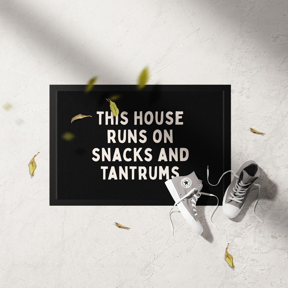 Doormat with text 'This house runs on snacks and tantrums' on a light gray floor with white sneakers and yellow leaves.