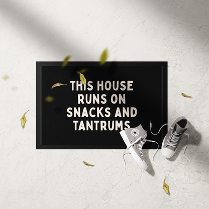 Doormat with text 'This house runs on snacks and tantrums' on a light gray floor with white sneakers and yellow leaves.