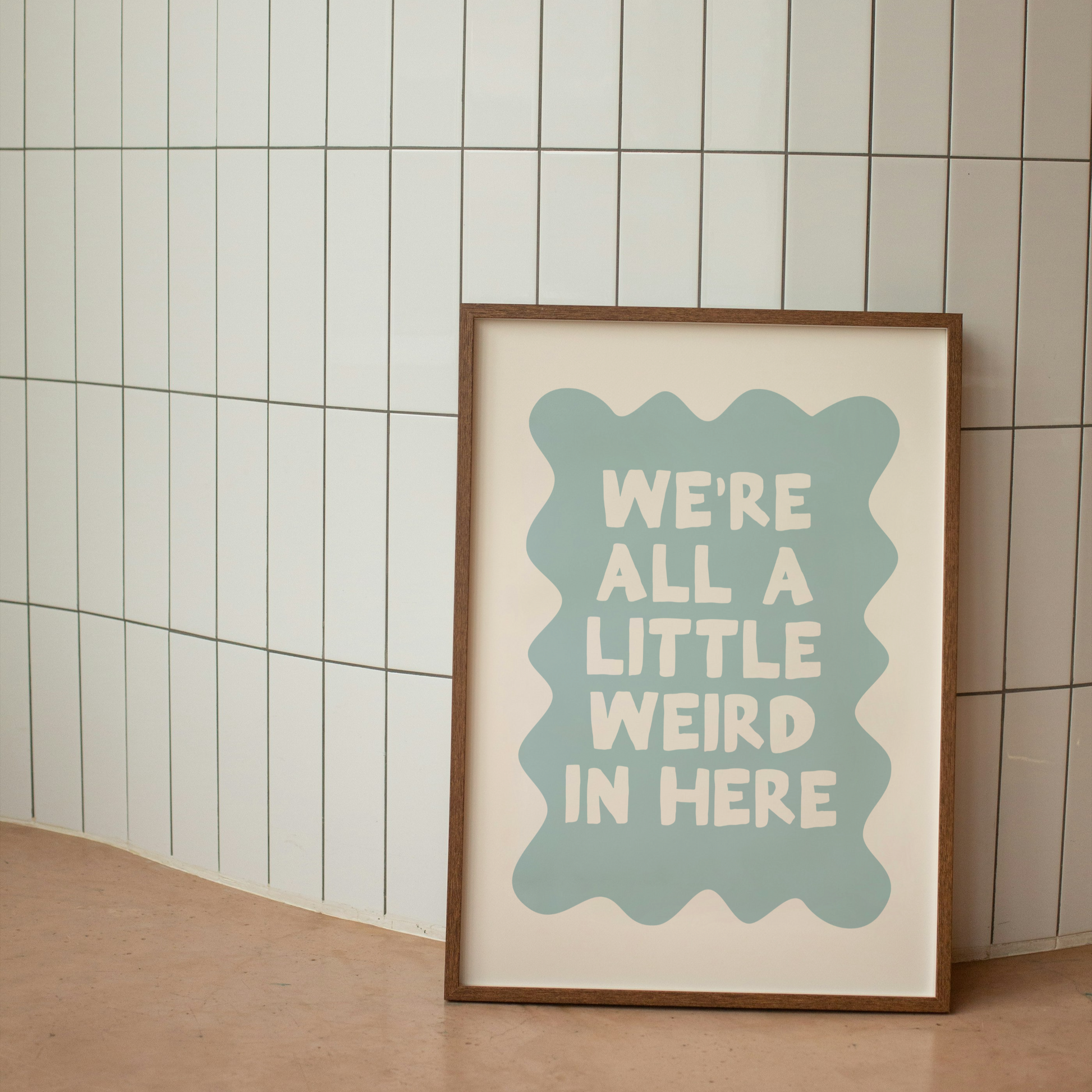 Framed poster with 'We're all a little weird in here' text on a tiled wall.